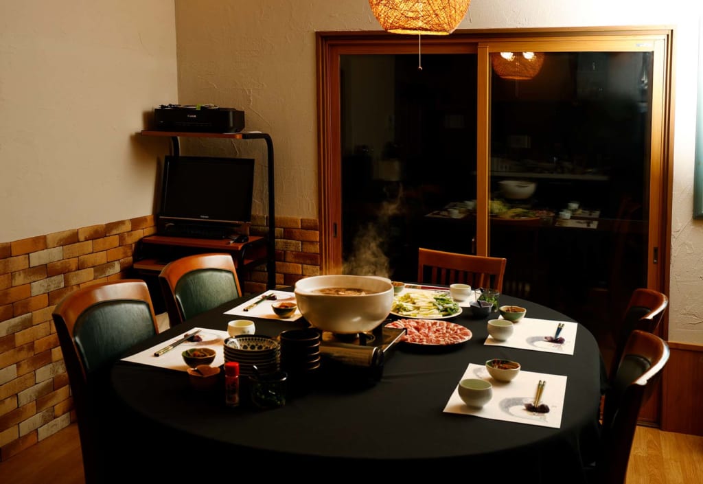 写真：夜、夕食が並べられたカフェスペースのテーブル。センターには湯気の立ちのぼる鍋と肉や野菜を盛り付けた大皿。